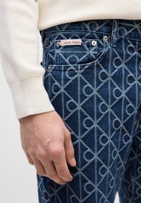 Vaqueros de mezclilla azul con un patrón geométrico de color azul claro, con una etiqueta blanca de Calvin Klein y herrajes plateados en los bolsillos.