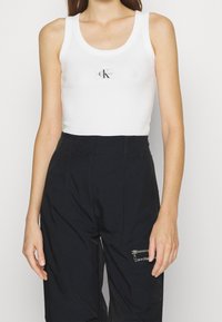 Vit ärmlös ribbad linne med en svart logotypapplikation i mitten, kombinerad med svarta höga byxor med zip-fickor.