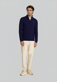 Muž stojící v námořnickém modrém svetr z zipem, bílém tričku pod ním, krémových kalhotách a béžových teniskách na pozadí s jednobarevným světlým pozadím.