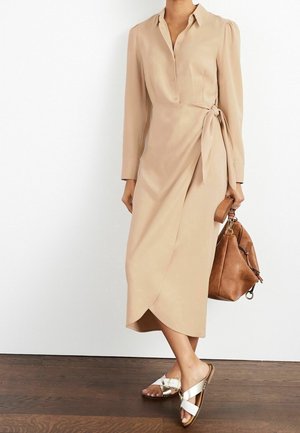 Abito beige a maniche lunghe con bottoni sul davanti e dettaglio con fiocco laterale. Indossato con una borsa a tracolla in pelle marrone e sandali bianchi a scivolo. Tessuto liscio.