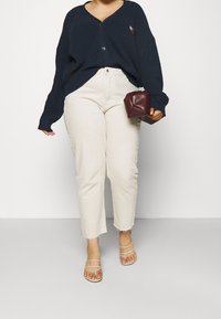 Granatowy prążkowany kardigan z zapięciem na guziki, kremowe zwężane spodnie z postrzępionym dołem oraz beżowe sandały na cienkich paskach, trzymająca bordową torebkę.