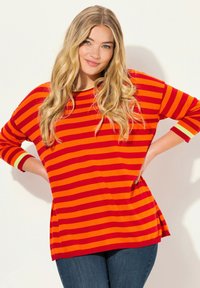 Röd och orange randig tröja med trekvartsärmar, ribbad halsringning och sidoklipp, kombinerad med mörkblå jeans.