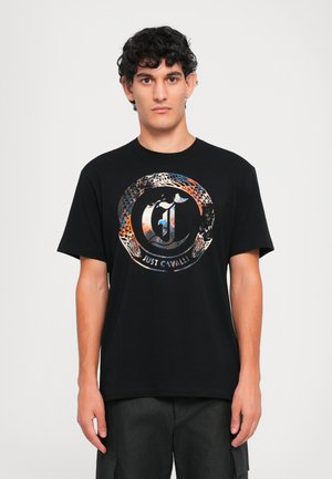 Jonge man met krullend haar, gekleed in een zwart T-shirt met een cirkelvormig, multikleurig logo en de tekst "Just Cavalli" aan de voorkant, staand tegen een effen achtergrond.