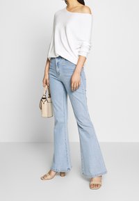 Femme portant un pull blanc à épaules dénudées, un jean évasé bleu clair, des sandales beiges et un sac à main crème texturé.