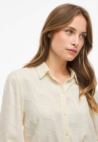 Blusa di colore crema con un motivo a onde testurizzato, colletto e chiusura con bottoni. Maniche lunghe con dettagli arricciati.