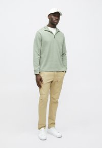 Light green, long-sleeved pullover with a zipper collar, paired with beige cargo pants. White sneakers and a white cap with a logo.