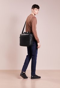 Svart läder axelväska med en strukturerad design. Har handtag upptill och en justerbar rem. Bärs med en brun tröja och mörka jeans.