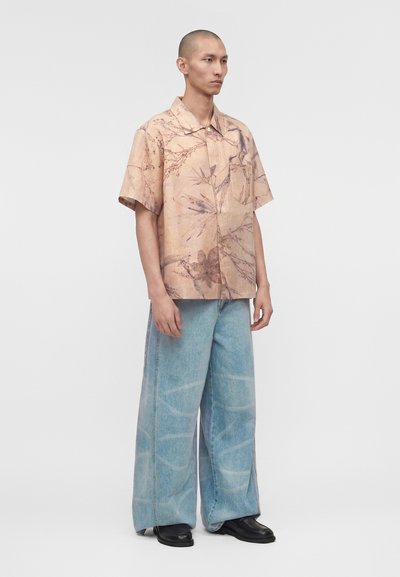 Άνδρας που στέκεται, φοράει μπεζ κοντομάνικο πουκάμισο με floral σχέδιο, φαρδιά ξεβαμμένα ανοιχτό μπλε τζιν και μαύρα παπούτσια σε απλό φωτεινό φόντο.