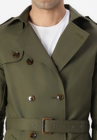 Homme portant un trench-coat croisé vert olive avec des boutons en écaille de tortue et une ceinture assortie sur un t-shirt blanc à col rond.