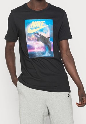 Schwarzes Baumwoll-T-Shirt mit einem bunten Wal-Graphic, Nike-Logo oben und Rundhals. Getragen mit hellgrauen Jogginghosen.