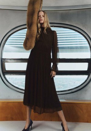 Vestido largo marrón con plisados y mangas transparentes, combinado con un estola de piel. La modelo está de pie junto a una gran ventana circular, llevando unos tacones negros.