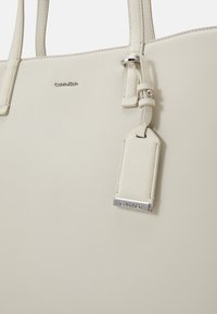 Borsa tote Calvin Klein in morbida pelle beige chiaro, con manici doppi, un'etichetta per bagagli testurizzata e hardware logo in argento. Finitura liscia.