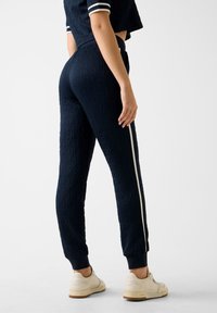 Eine Frau trägt navyblaue, strukturierte Jogginghosen mit einem weißen Seitenstreifen, ein passendes kurzes Oberteil und beige Sneakers und steht vor einem weißen Hintergrund.