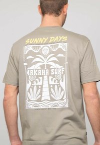 T-shirt vert olive avec un design imprimé blanc représentant un palmier, un soleil, et le texte "JOURS ENSOLEILLÉS" et "MAKAHA SURF" dans un style rétro.