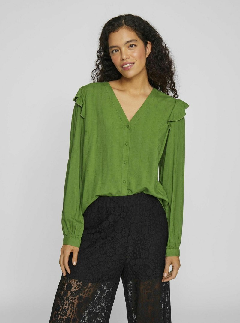 Green button-up blouse with a V-neck, ruffled shoulder accents, and long sleeves, paired with black lace pants featuring floral patterns.