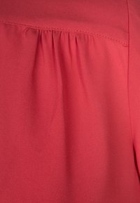 Červená, lehká tkanina s mírně strukturovaným povrchem. Obsahuje nařasený detail v oblasti výstřihu a hladký, splývavý siluetu.