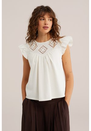 Femme aux cheveux bouclés portant un chemisier blanc à motifs carrés ajourés et manches à volants, associé à un pantalon plissé marron foncé.