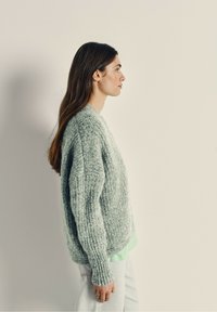 Overmaat, mintgroene, gebreide trui met een textuurafwerking en ribbelmanchetten, gecombineerd met lichte spijkerbroek. Zijaanzicht.