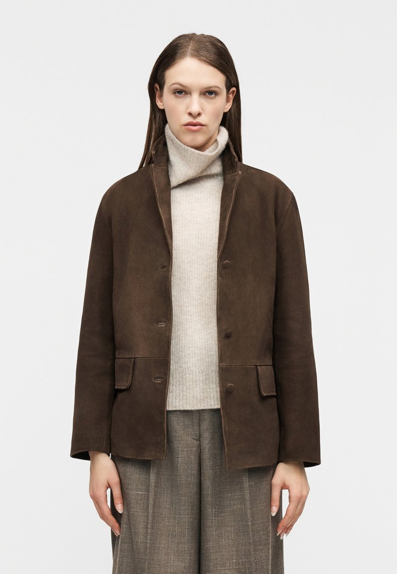 Γυναίκα που φοράει καφέ σουέντ μπο υφάν πάνω από μπεζ πουλόβερ με λαιμό, και γκρι καρό παντελόνι φαρδύ, στέκεται μπροστά σε απλό φωτεινό φόντο.