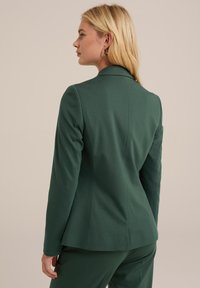 Blazer su misura verde con una vestibilità strutturata, revers a notches e cuciture delicate. Realizzato in un tessuto liscio per un aspetto elegante.