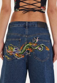 Dark blue denim jeans featuring vibrant dragon embroidery on the back pockets, with a textured surface and traditional five-pocket design.