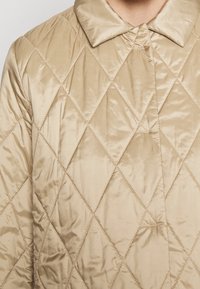 Quilted jacket in beige satin with a diamond pattern, featuring a folded collar and a smooth texture. Design is simple, minimalistic, and functional.