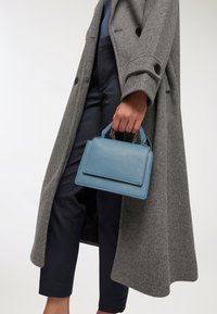 Jasnoniebieska skórzana torebka o prostokątnym kształcie, z małymi uchwytami i łańcuszkowymi akcentami, trzymana w ręce ubranej w szary płaszcz na granatowe spodnie.
