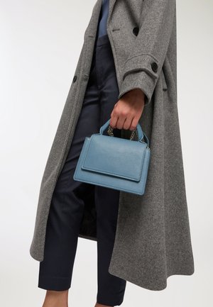 Borsa a mano in pelle azzurro chiaro di forma rettangolare, con manici piccoli e dettagli in catena, tenuta in una mano che indossa un cappotto grigio sopra pantaloni blu navy.