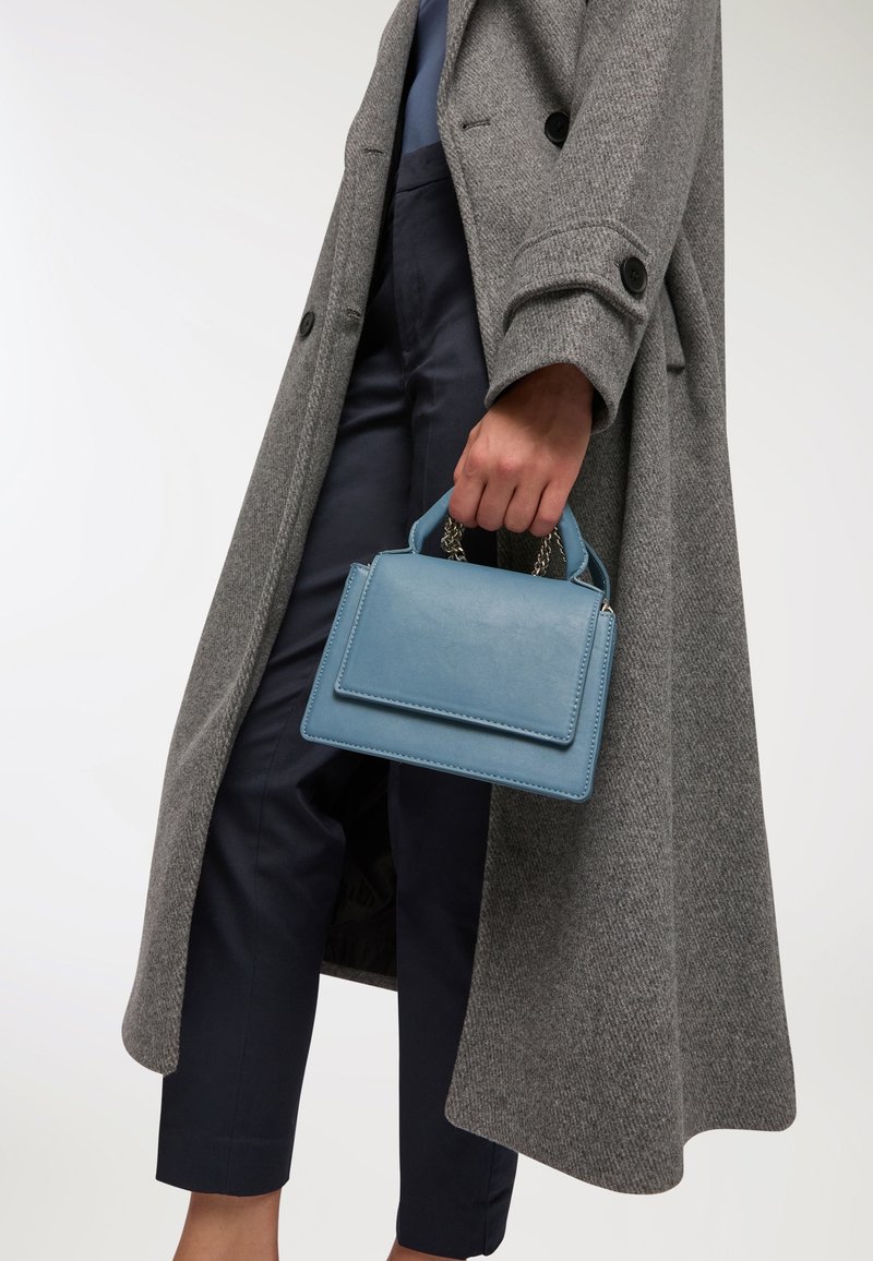 Jasnoniebieska skórzana torebka o prostokątnym kształcie, z małymi uchwytami i łańcuszkowymi akcentami, trzymana w ręce ubranej w szary płaszcz na granatowe spodnie.