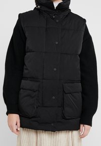 Chaleco acolchado negro con cuello alto, botones a presión y dos bolsillos frontales. La textura suave y el diseño acolchado acentúan el calor.