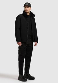 Zwarte pufferjack met ritsafsluiting en bontkraag, gecombineerd met zwarte cargo-broeken en stevige zwarte laarzen. Accessoires zijn een pet.