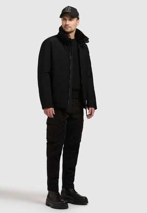 Mustat untuvapusero, jossa on vetoketjukiinnitys ja turkiskaulus, yhdistettynä mustiin cargo-housuihin ja koviin mustiin saappaisiin. Asusteena on lippis.