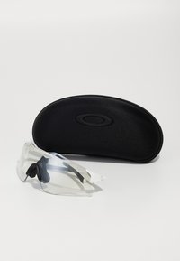 Klare Sportsonnenbrille mit einem weißen Rahmen und schwarzen Gummibezügen, neben einem schwarzen ovalen Schutzetui mit erhabenem Logo.