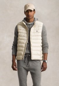 Krēms krāsas puffa veste ar augstu apkakli, uzvilkta pār pelēku džemperi ar kapuci, saskaņota ar pelēkiem sporta biksēm. Bežs vāciņš un rokas pulkstenis papildina izskatu.