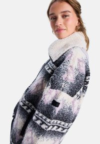 Giacca in pile con colletto alto, motivi geometrici grigi, bianchi e rosa, tessuto testurizzato e tasca laterale con zip.