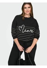 Femme portant un pull noir à rayures blanches avec le texte « love », tenant un sac à main noir, debout devant un fond clair.