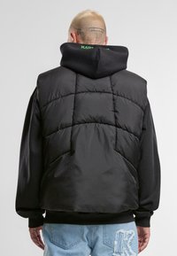 Crni napuhani prsluk s quilted uzorkom i visokim ovratnikom, nošen preko crne dukserice. Ima opušteni kroj i rebraste manžete.