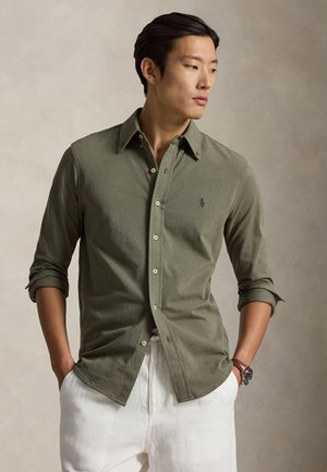 Homme aux cheveux foncés portant une chemise olive à manches longues boutonnée et un pantalon blanc, mains dans les poches, regardant sur le côté.