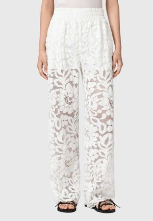 Kvinde iført hvide, vide blondebukser med blomsterprint og sorte sandaler, stående foran en ensfarvet baggrund.