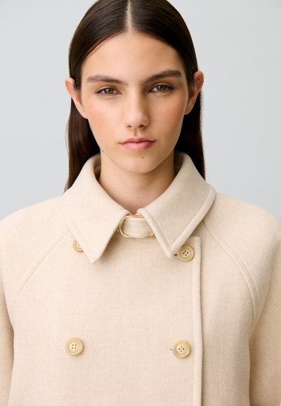 Giovane donna con capelli scuri e lisci indossa un cappotto beige doppiopetto con ampio colletto e dettagli con bottoni su sfondo semplice.