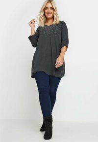 Donkergrijze tuniek met V-hals, met een versierde halslijn met kleine glanzende details. Gecombineerd met donkerblauwe leggings en zwarte enkellaarsjes.