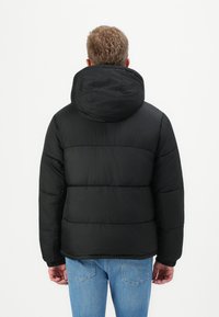 Schwarze Pufferjacke mit Kapuze, mit gesteppte Abschnitten und glatter Textur. Getragen mit hellblauen Jeans, von hinten gesehen.