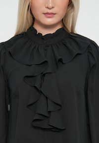 Black blouse with large ruffled collar, layered front ruffles, and button closure. Made of smooth fabric, features a sleek silhouette.