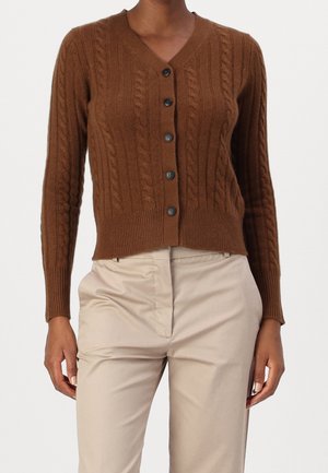 Cárdigan marrón de punto con botones, combinado con pantalones beige de cintura alta, en una persona de pie contra un fondo blanco.