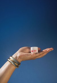 Hvit beholder med rød skrift, merket "EGYPTIAN MAGIC All Purpose Skin Cream," holdt i en hånd. Glatt tekstur, sirkulær form. Gullarmbånd på håndleddet.