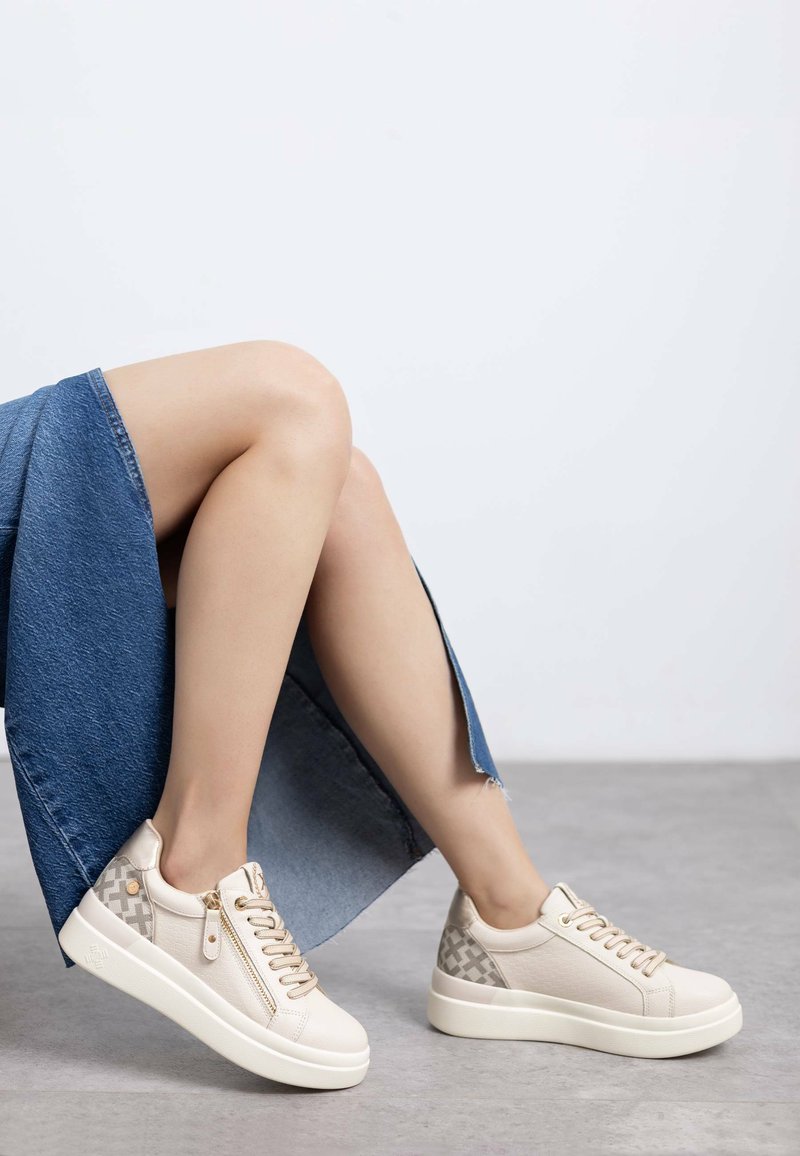 Zapatillas beige con lados texturizados, cordones en la parte delantera y detalle de cremallera. Presentan una suela blanca gruesa y un acabado suave. Se llevan con una falda de mezclilla.