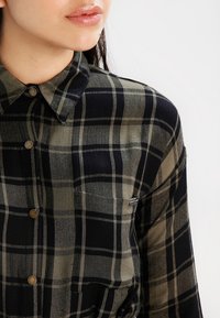 Chemise à carreaux verte et noire, boutonnière avec un col pointu, dotée de boutons métalliques et d'une poche sur le côté gauche. Texture de tissu douce.