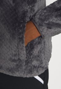 Grå fleecejacka med en mjuk, texturerad yta, med en dold ficka och kontrasterande bruna detaljer vid handlederna.