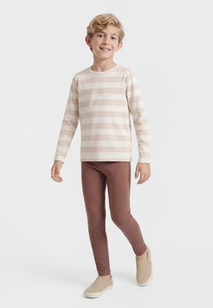 Boy standing and smiling, wearing beige and white striped long-sleeve shirt, brown pants, and beige slip-on shoes on white background.