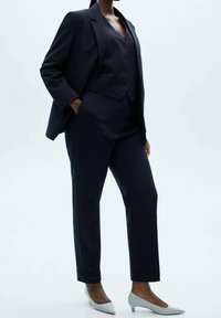 Námornícky oblek na mieru s dobře padnoucím sakem s klopami, odpovídajícími kalhotami a vestou na tři knoflíky, doplněný světle šedými špičatými podpatky.